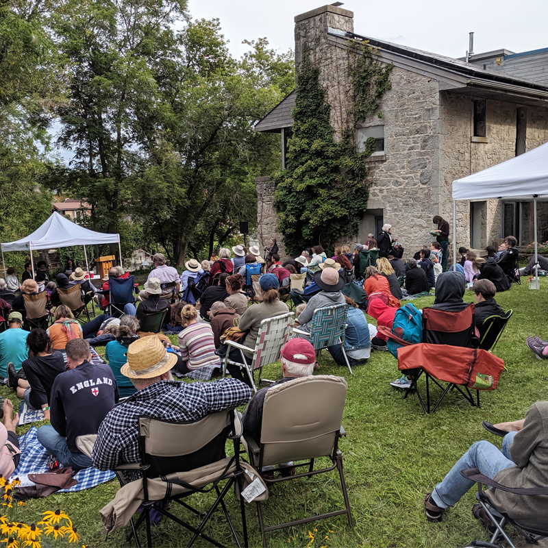 Photo of a packed audience sitting in lawn chairs beside a heritage home. 