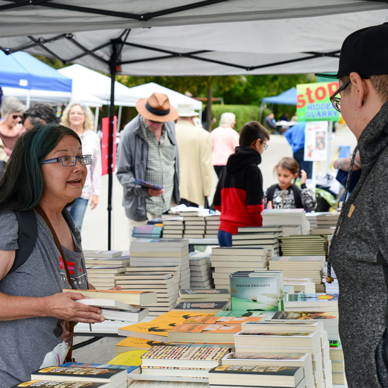 Photo of two individuals chatting over a table stacked with books.
